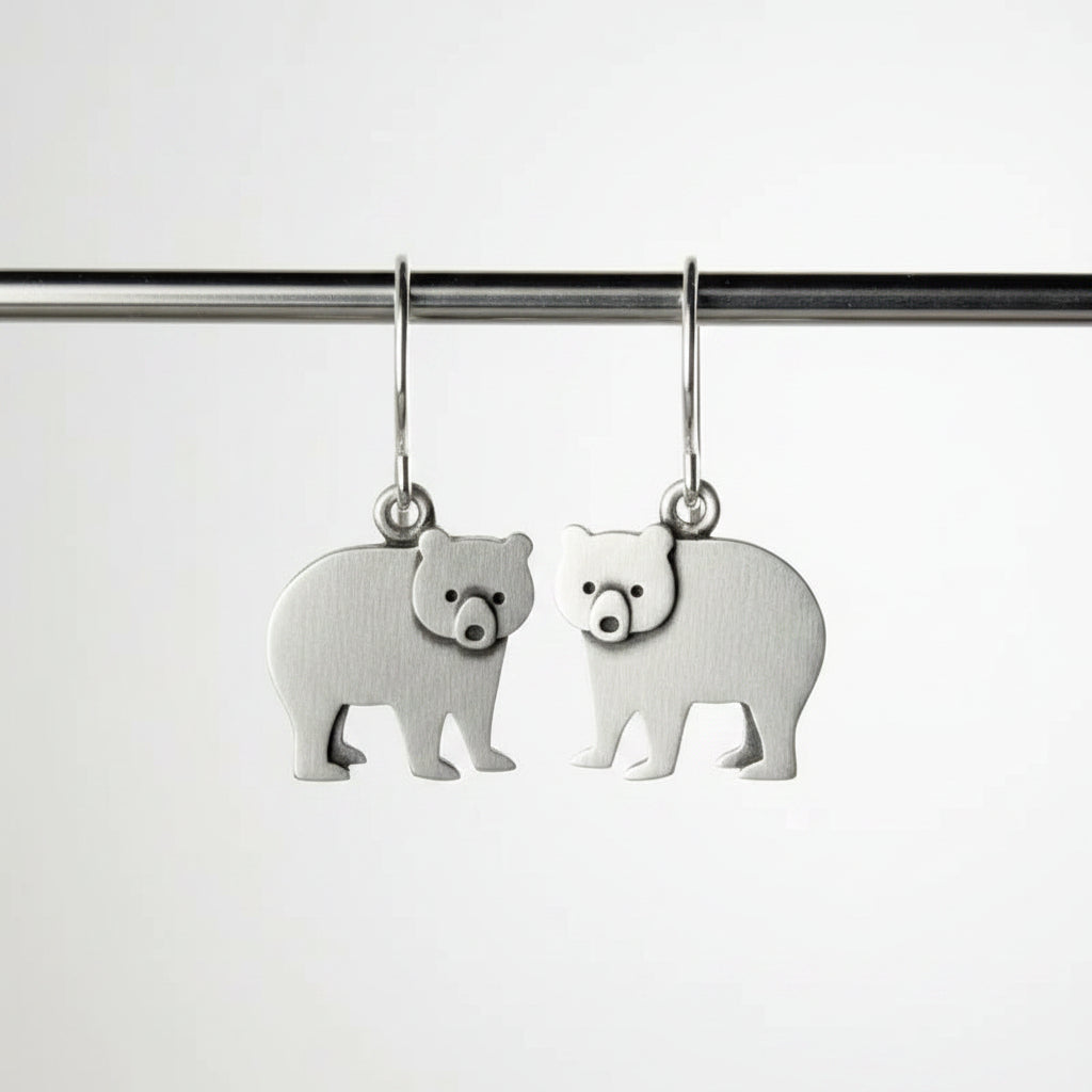 Silver bear-shaped earrings on a hook, held by a finger against a neutral background.